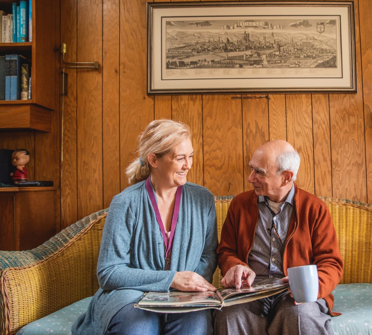 An older malw looking at an album while sitting on a couch inside the living room with his younger female carer