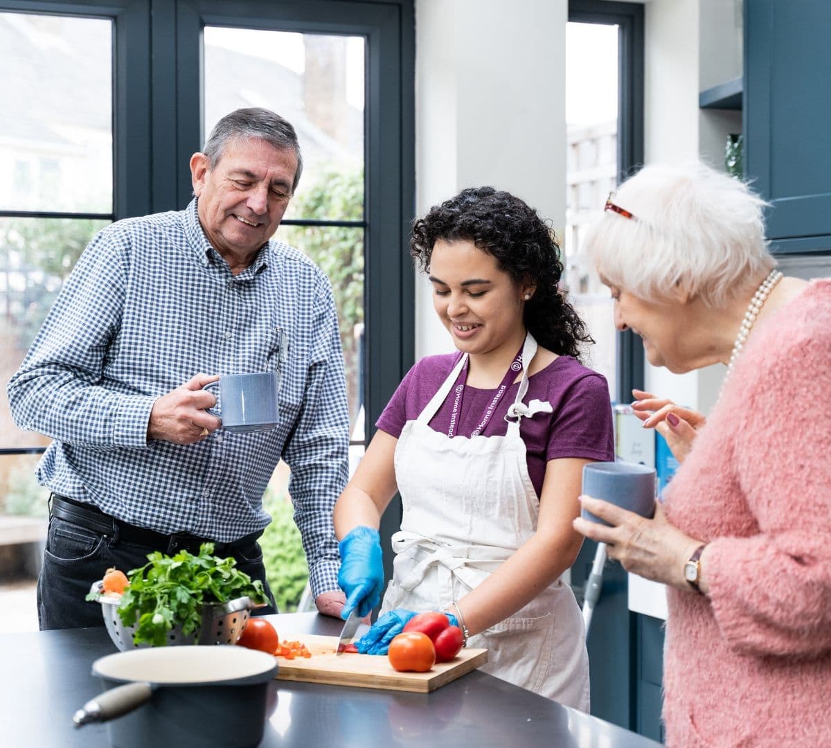 Three people in a kitchen, smiling as one chops vegetables while the others watch and hold mugs. - Home Instead