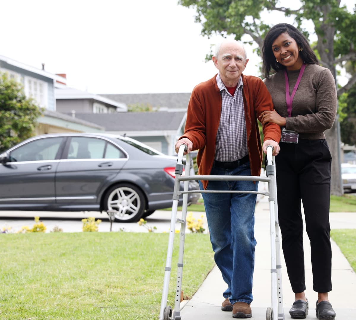 Male older adult walking outdoors while using a walker and with grey hair and wearing a sweater while being assisted by his younger female carer with black hair both smiling