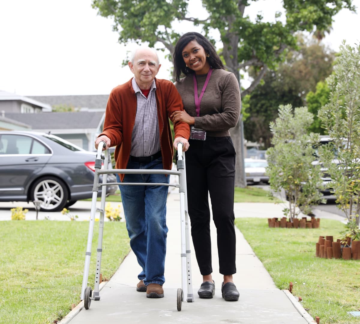 An older woman with short hair and wearing pink walking outdoors with her younger carer with blonde hair wearing printed black both happy and smiling while walking and surrounded by green grass