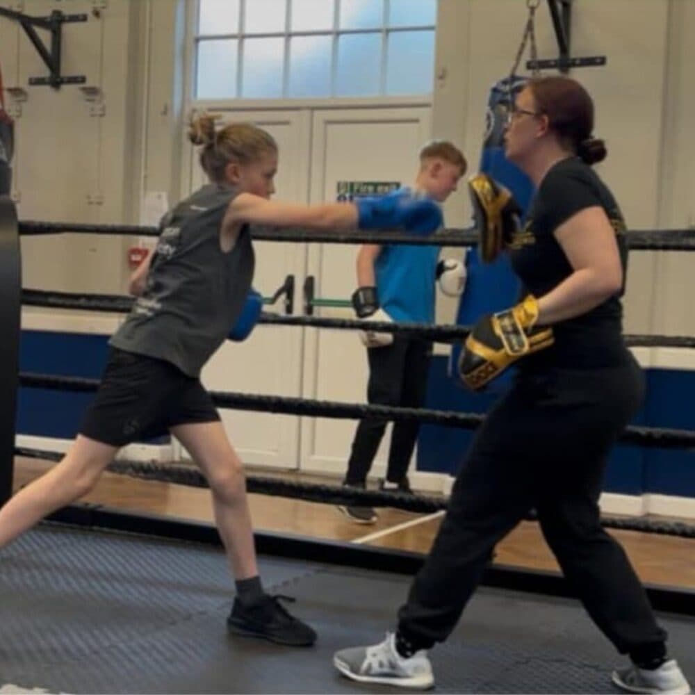A young girl in boxing gear punches mitts held by her coach in a boxing gym. - Home Instead