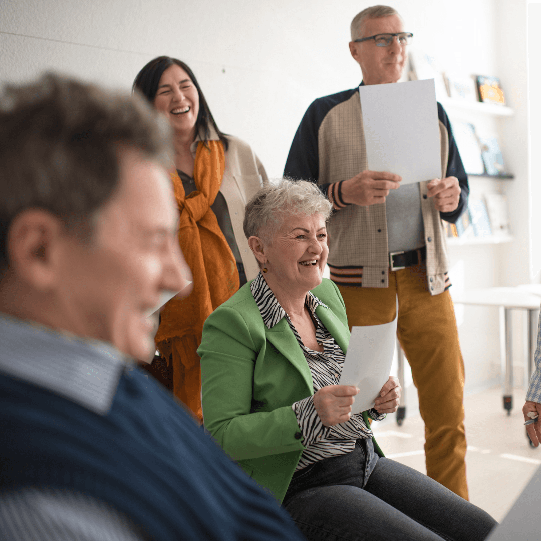 A group of adults smiling and holding song sheets during a relaxed group singing session.