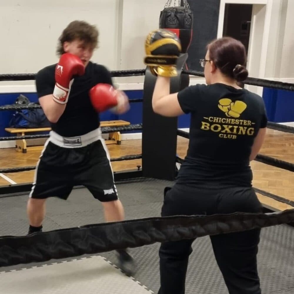 A boxer trains in a ring, punching pads held by a coach wearing a "Chichester Boxing Club" shirt. - Home Instead