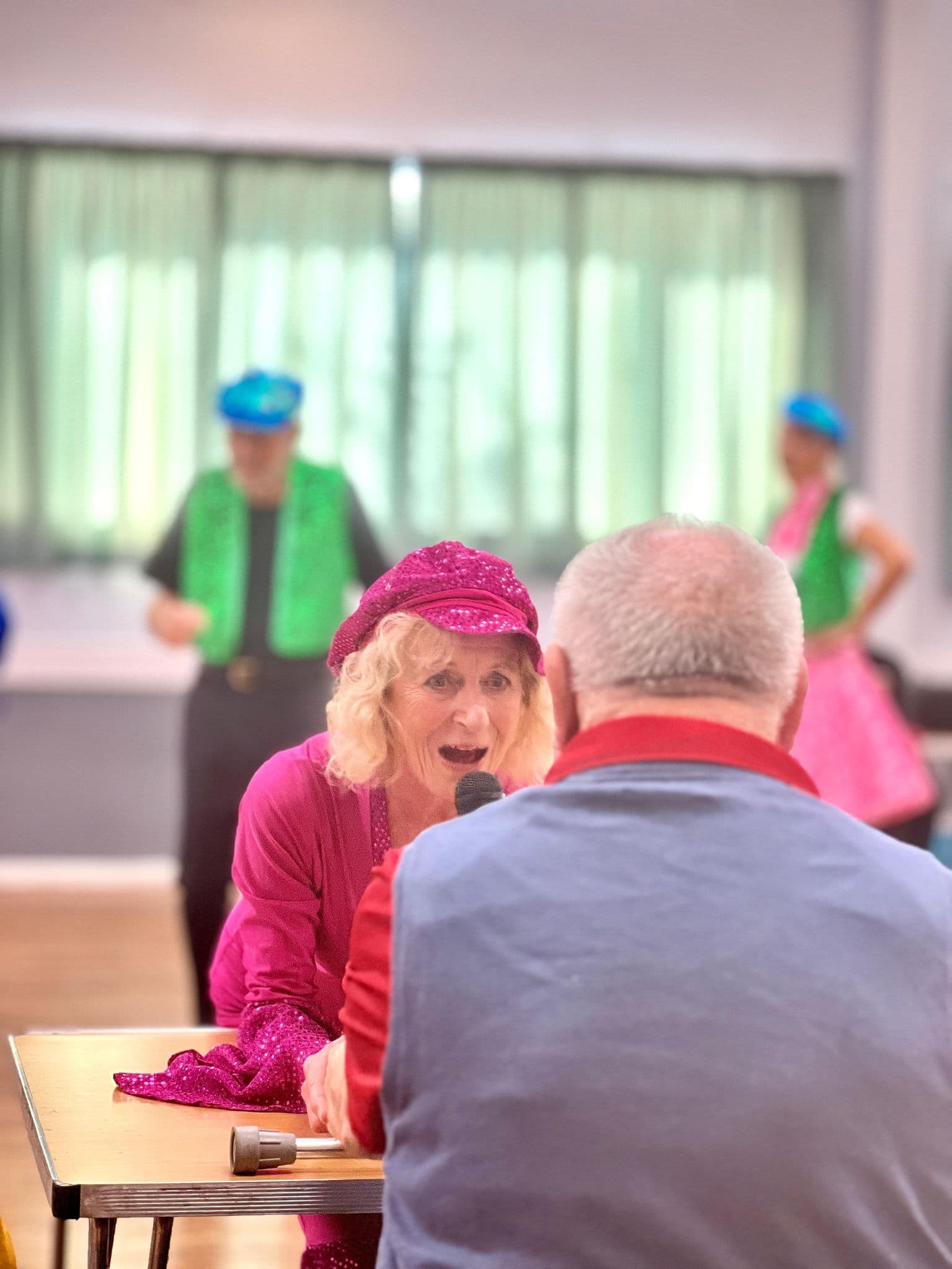 A woman in bright pink sings into a microphone facing an older man, with performers blurred in the background. - Home Instead