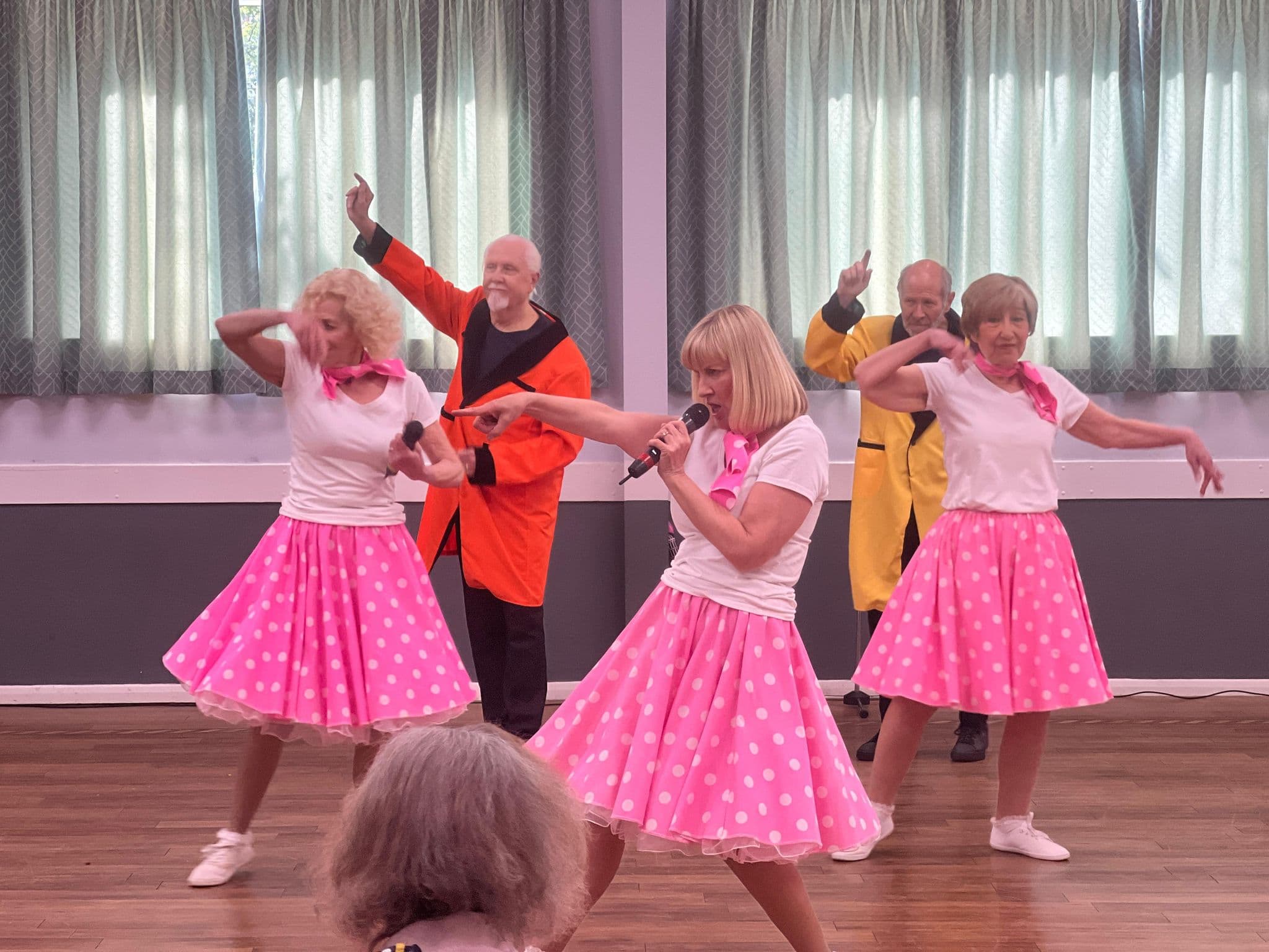 Three women in pink polka dot skirts and two men dance and sing on a stage with microphones. - Home Instead