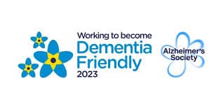 Logo for Alzheimer’s Society with the text "Working to become Dementia Friendly 2023" and blue flowers. - Home Instead