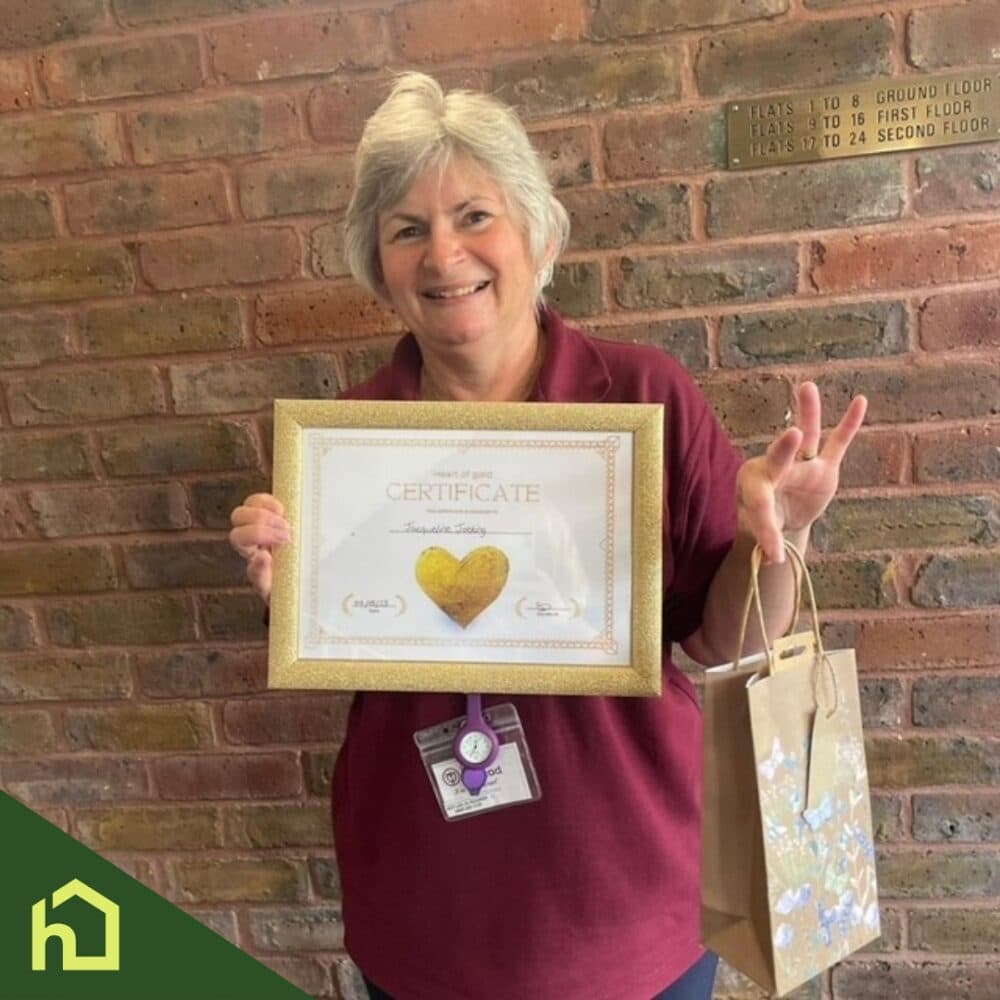 Smiling woman holding a certificate and gift bag, standing by a brick wall, wearing a maroon shirt. - Home Instead