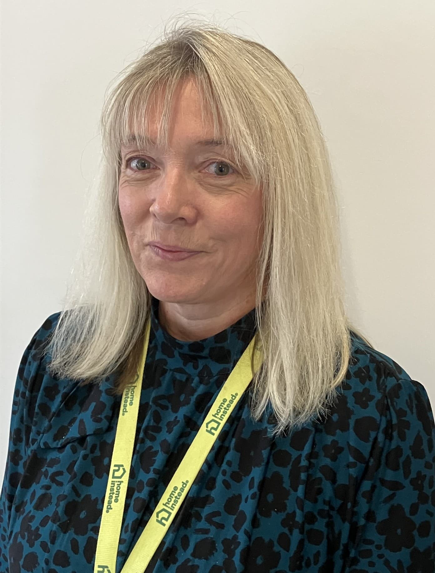 A woman with long blonde hair wearing a printed dress and a lanyard stands in front of a light-colored wall. - Home Instead