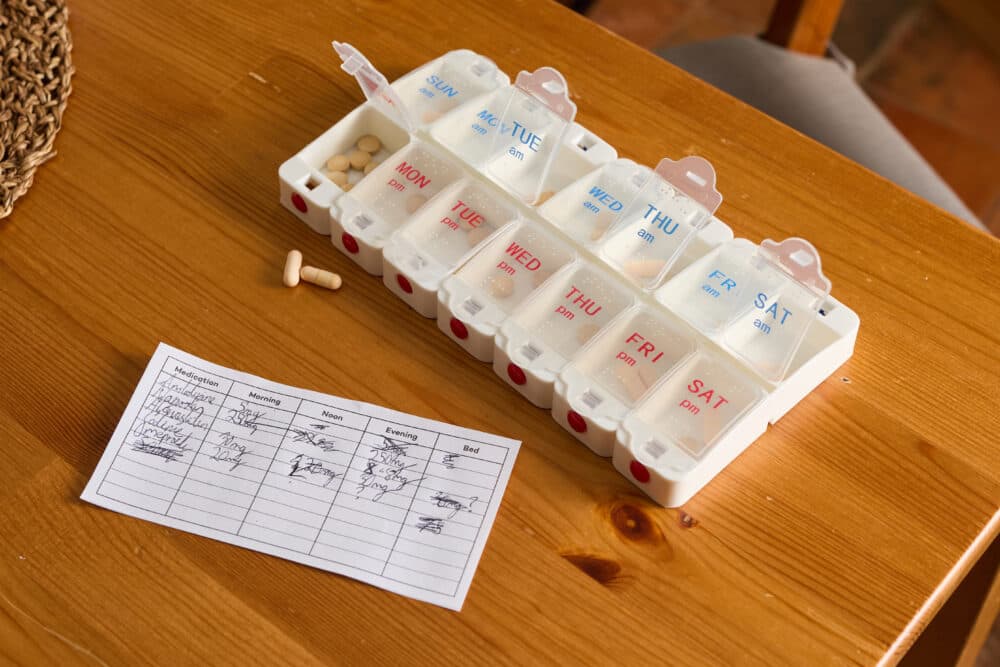 A weekly pill organizer and a medication schedule on a wooden table, with some pills next to them. - Home Instead