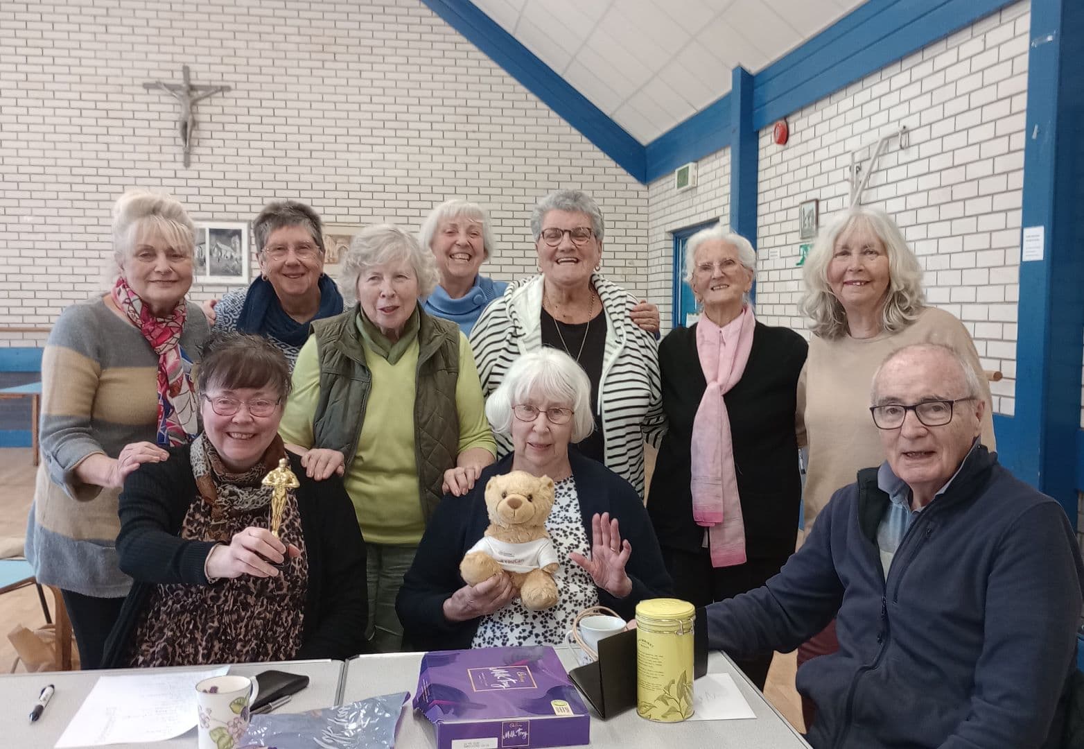 A group of smiling older adults poses indoors at a table with tea, sweets, and a teddy bear. - Home Instead