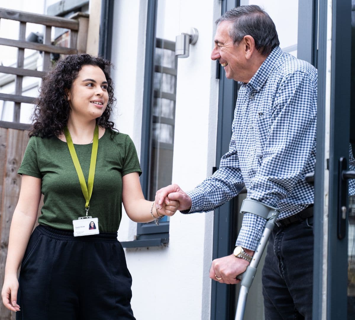 Young woman with a name badge helps an older man on crutches at a doorway, smiling at each other. - Home Instead