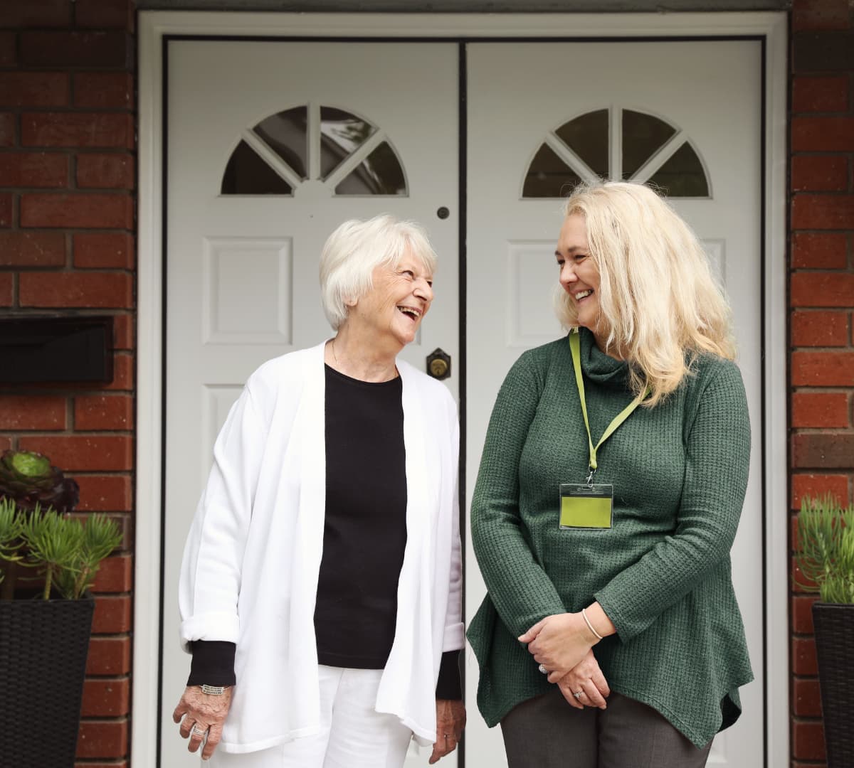 An older female adult wearing black and white and with short hair with her younger female carer with long blonde hair and wearing green both in front of the door and happy and smiling