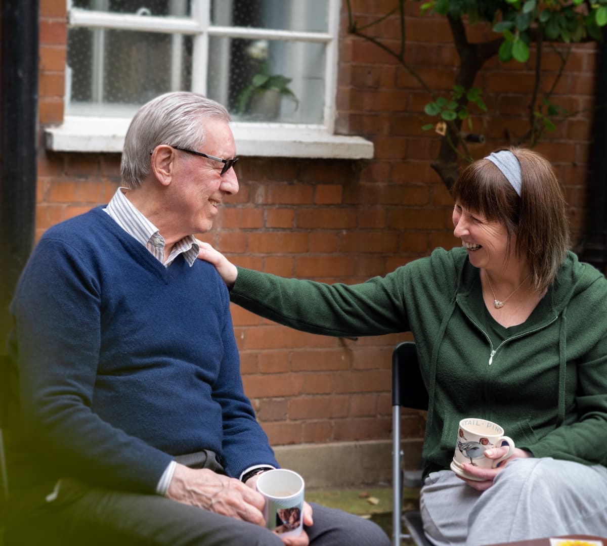 An older man with grey hair and wearing eyeglasses and blue sweater having coffee with his younger female carer having coffee outside the house