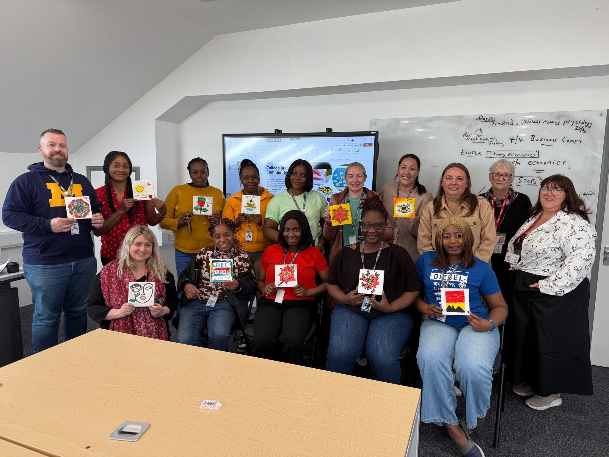 A group of people smiling and holding up colorful artwork in a classroom setting. - Home Instead