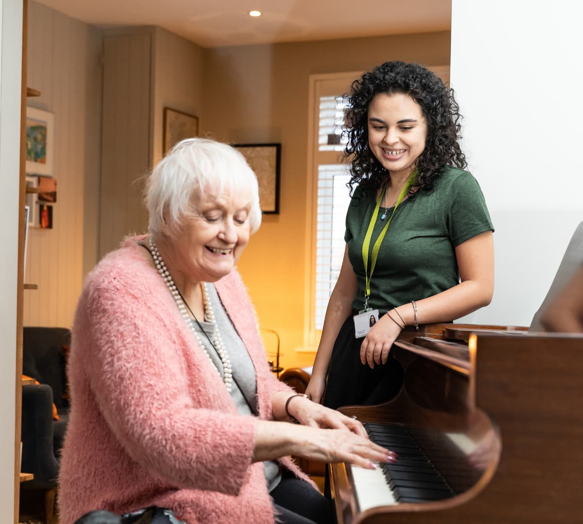 An older woman with short white hair and wearing pink while playing the piano with her younger female carer with black curly hair adn wearing reen shirt both happy and smiling inside the house
