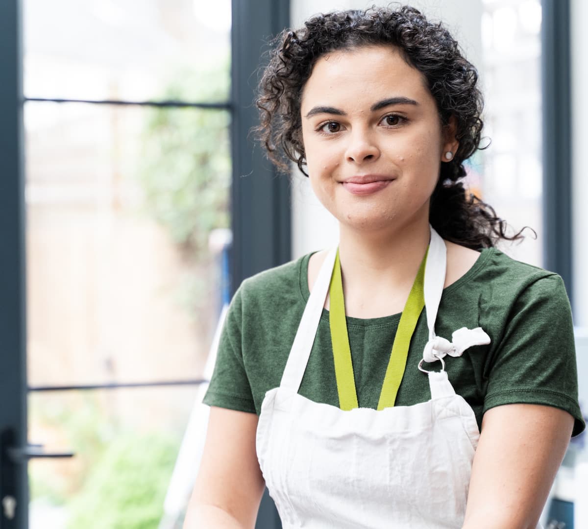 A female carer with long curly hair wearing green and white apron smiling while inside the kitchen