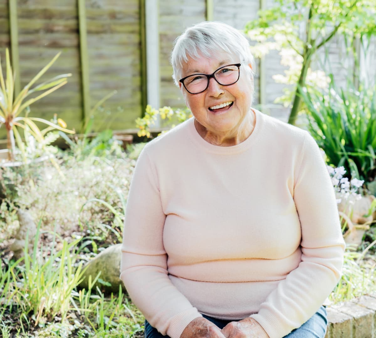 An older woman with grey hair smiling and wearing eyeglasses while surrounded with plants in the garden