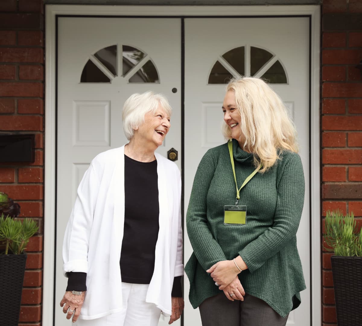 Older female adult with white hair and wearing whote with her younger female carer with long blonde hair wearing green both happy and smiling at the front door of the house