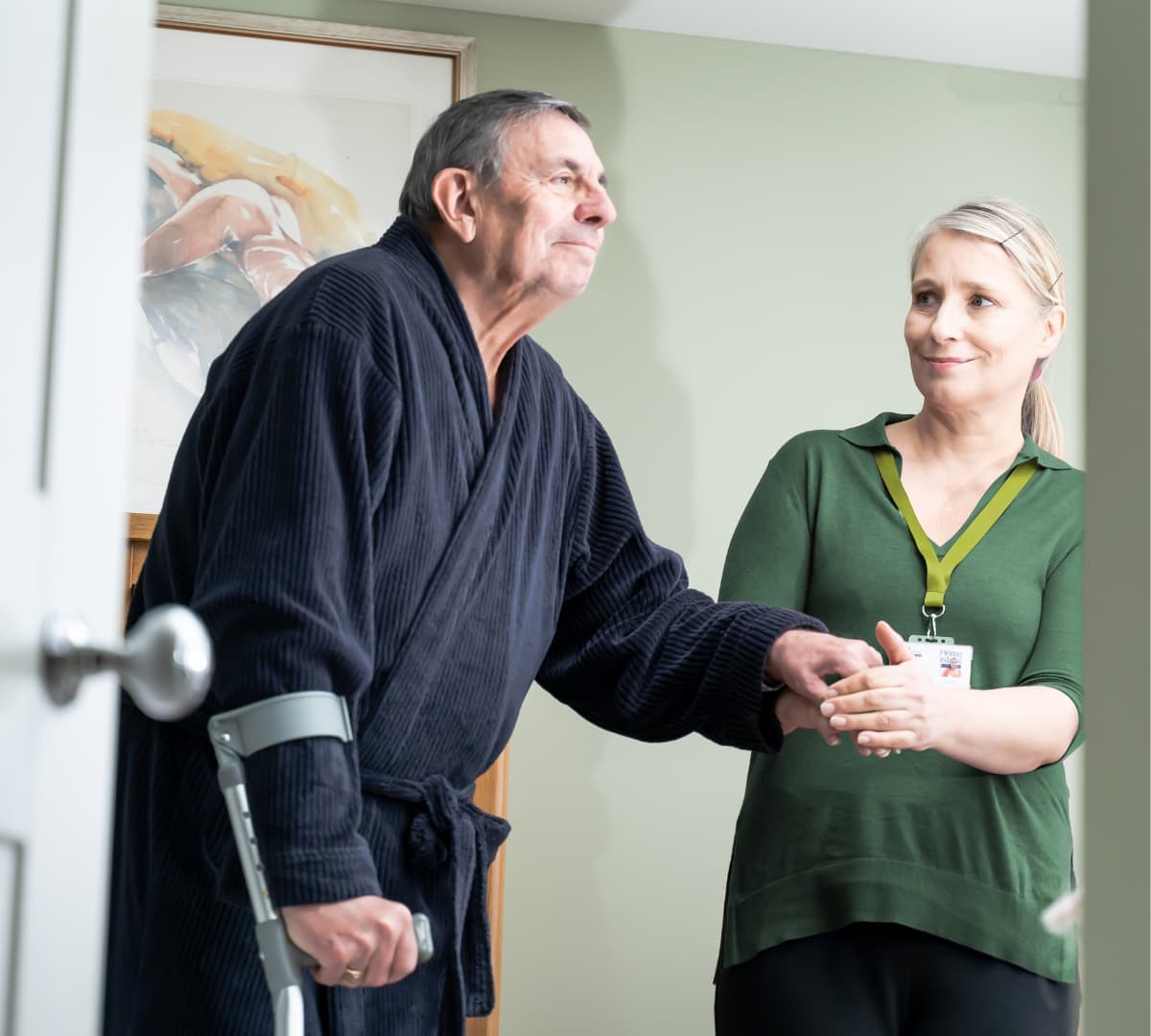 An older male adult with grey and wearing a bathrobe walking inside the house while using a crutch and being helped by his younger female carer wearing green and smiling