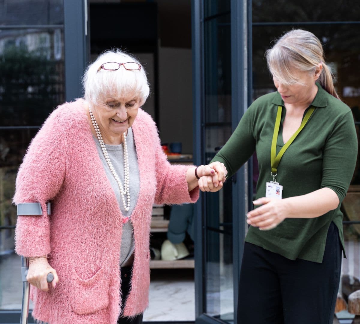 An older female adult with white hair and wearing pink and using a crutch walking outside while being helped y her younger female carer wearing green
