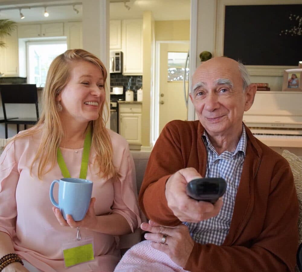 An older man watching tv while holding the remote control with his younger female carer with long blonde hair and wearing pink while holding a cup inside the home both happy and smiling