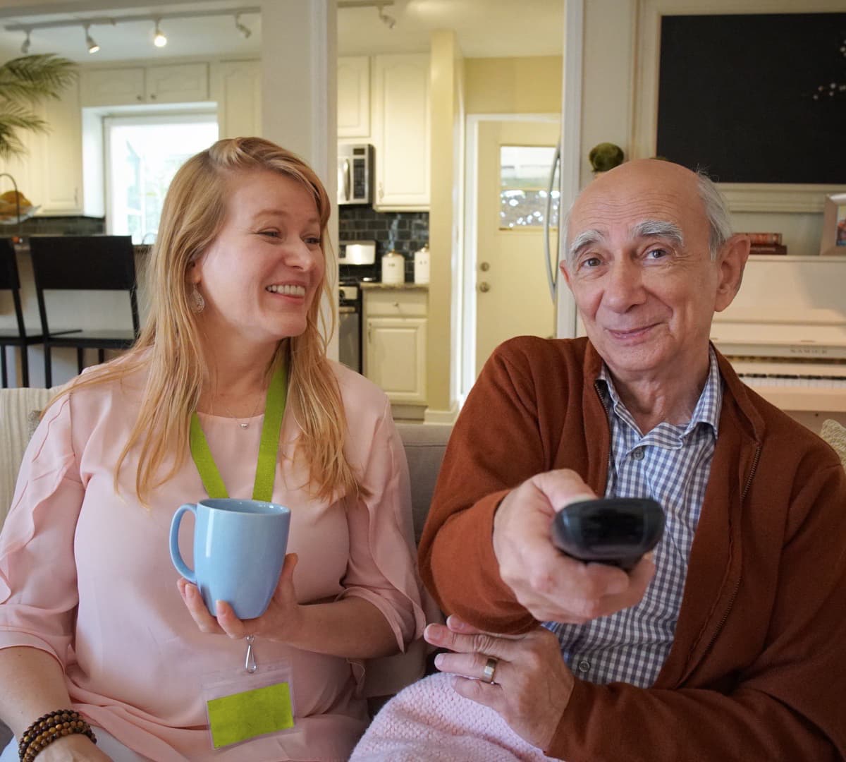Older male adult with grey hair and wearing a sweater holding the television remote control with his female carer wearing pink with long blonder hair holding a cup of coffee inside the living room both happy and smiling