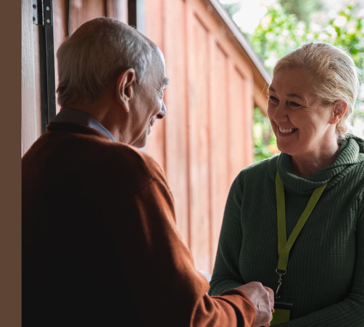 An older man welcoming his younger female carer wearing green at the door of his house