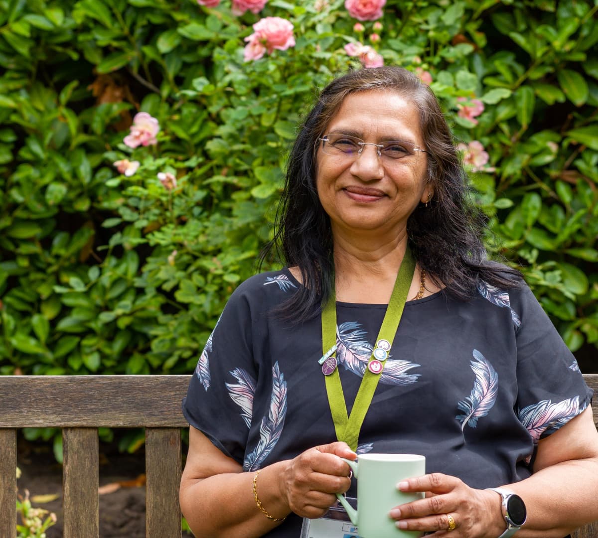 A woman with long black hair wearing eyeglasses and sitting on a bench and holding a cup surrounded by green plants in the garden