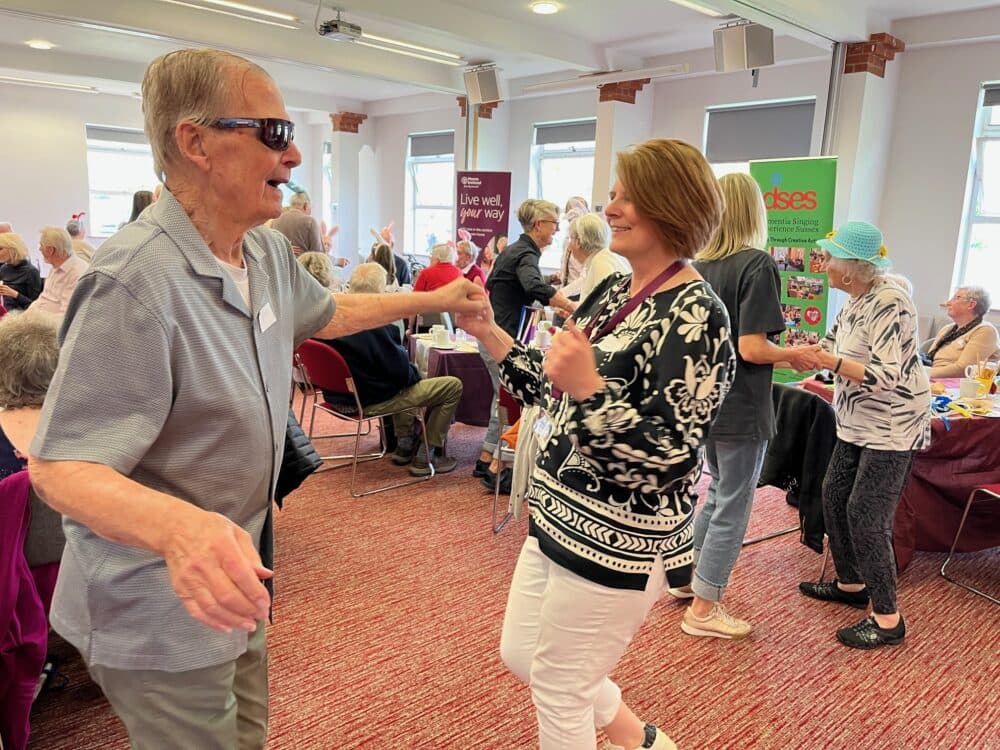 An older man and a woman dance together, smiling, at a lively community event in a bright room. - Home Instead