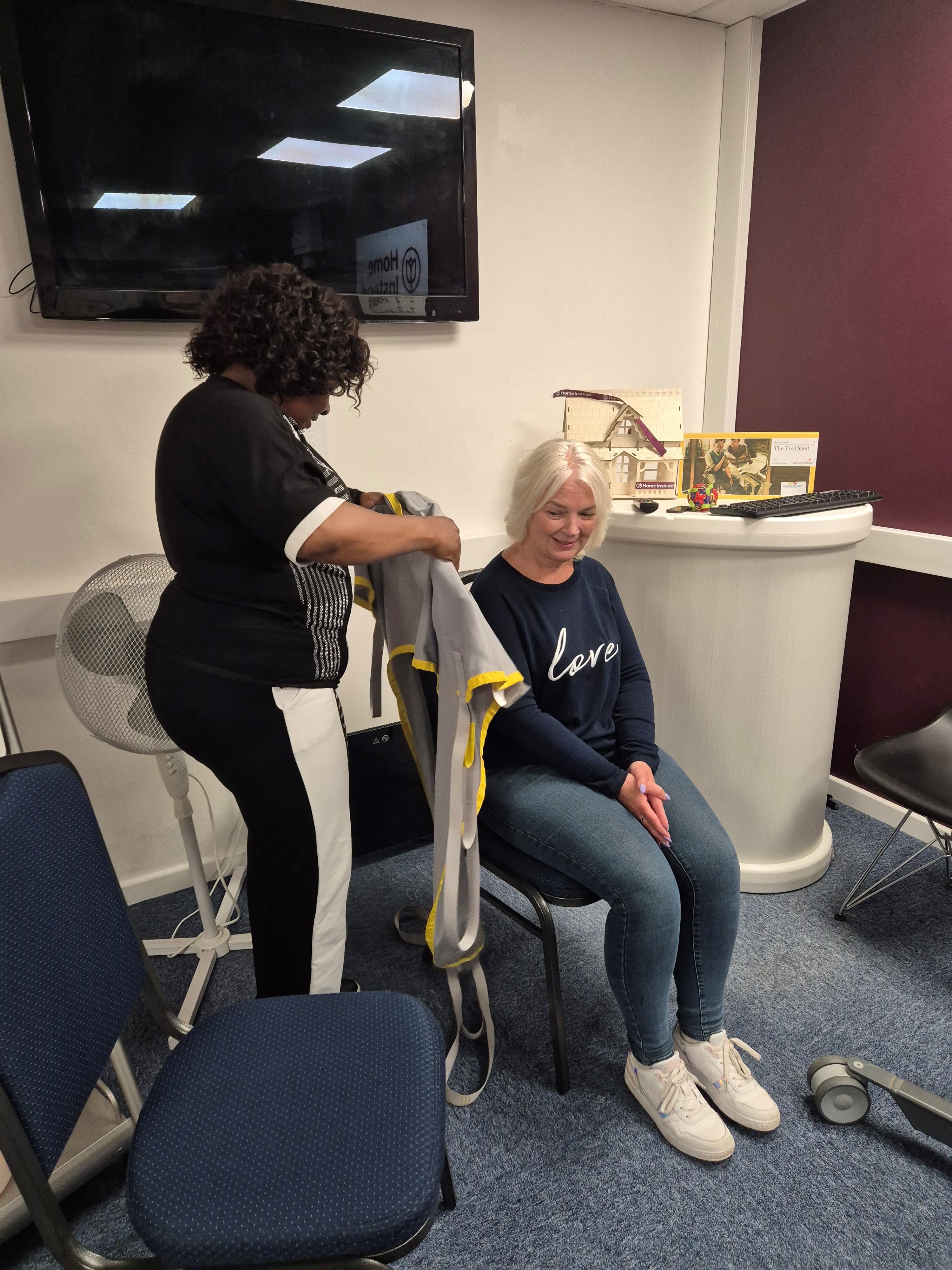 Deborah fits a grey and yellow sling around Daisy’s torso while Daisy sits smiling on a chair.