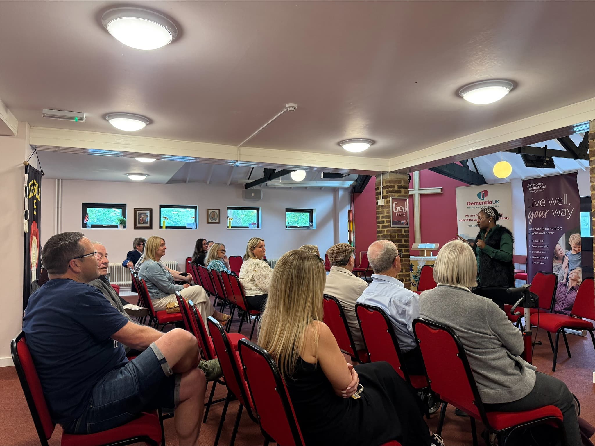 People seated on red chairs in a community room, listening to a speaker at the front near banners and a cross. - Home Instead