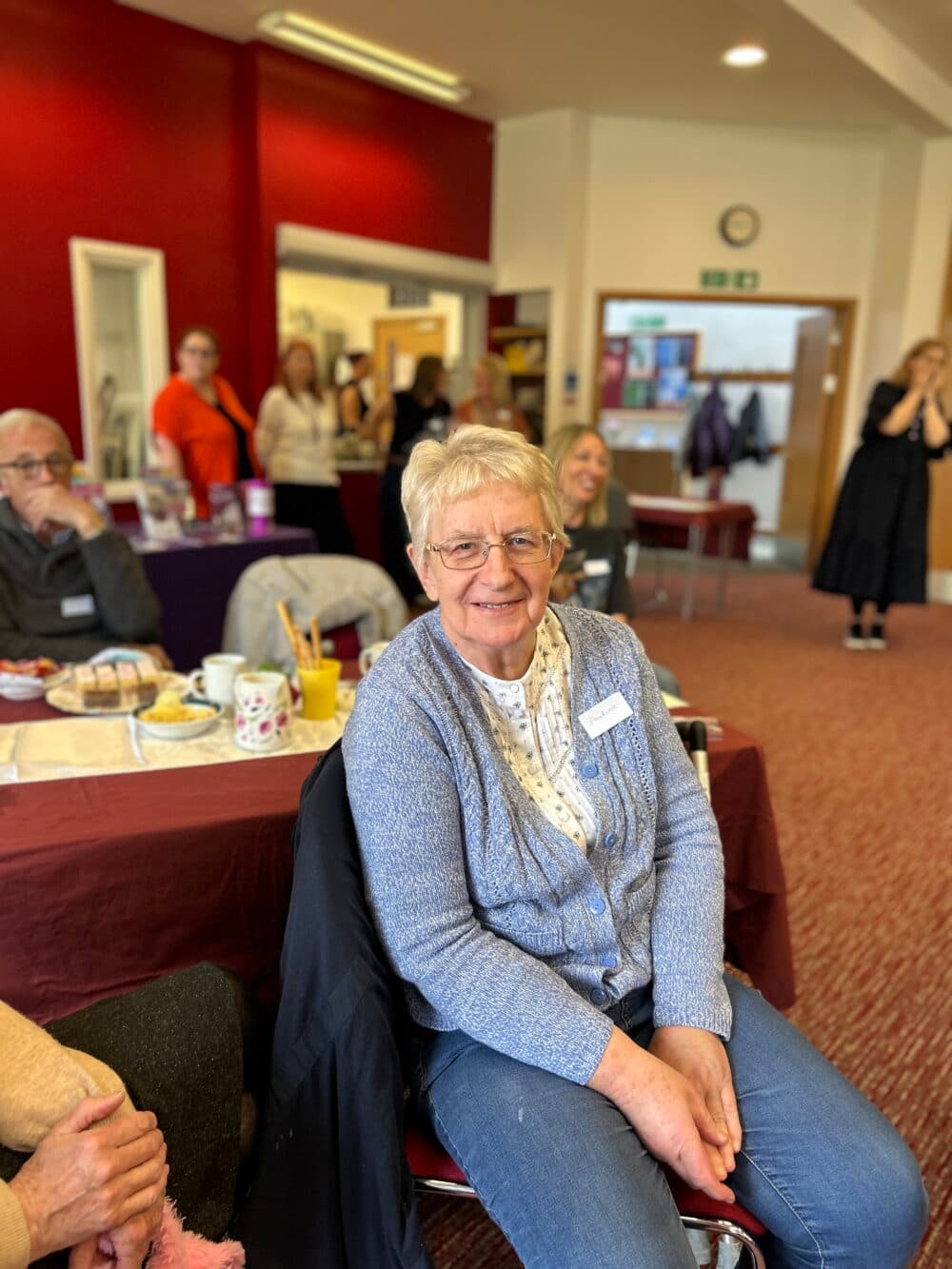 An older woman smiles while sitting in a bright room with people and refreshments in the background. - Home Instead
