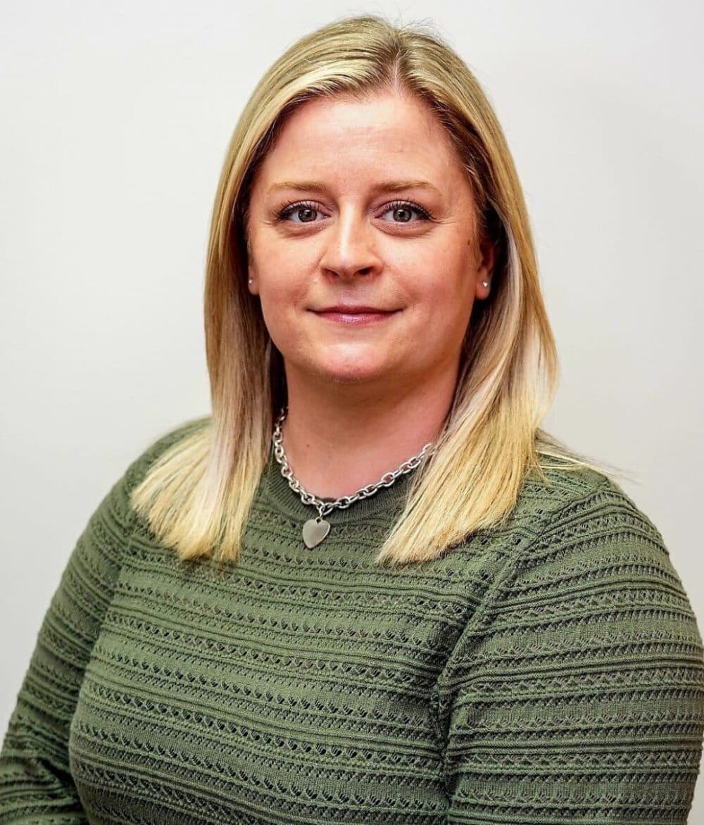 Woman with straight blonde hair, wearing a green textured sweater and silver necklace, smiling slightly. - Home Instead