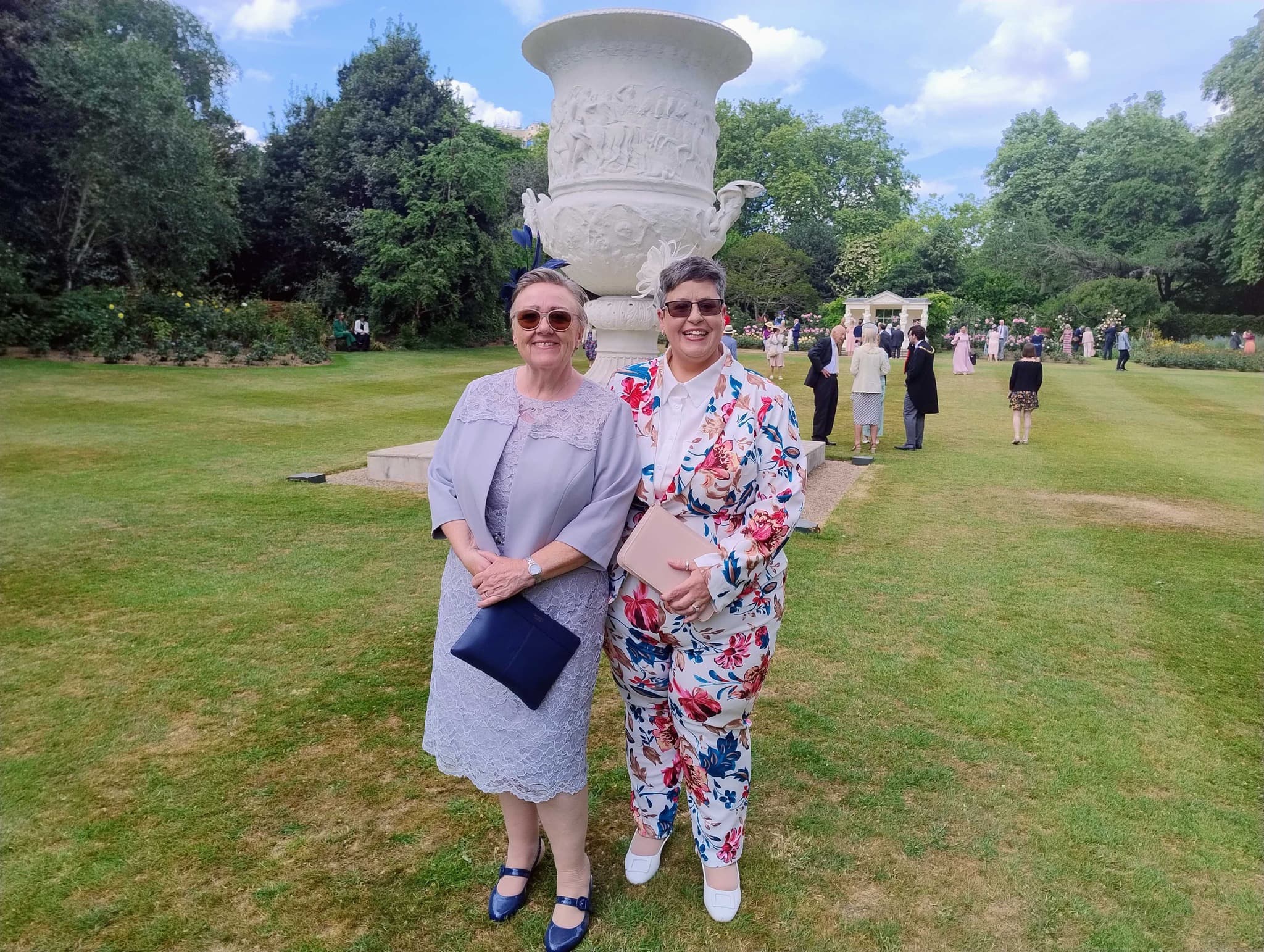 Two women posing in front of a large decorative urn in a garden, dressed in formal outfits on a sunny day. - Home Instead