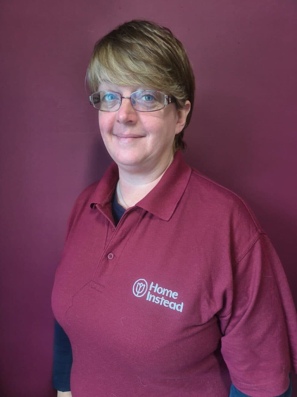 Person wearing glasses and a burgundy "Home Instead" polo shirt standing in front of a matching wall. - Home Instead