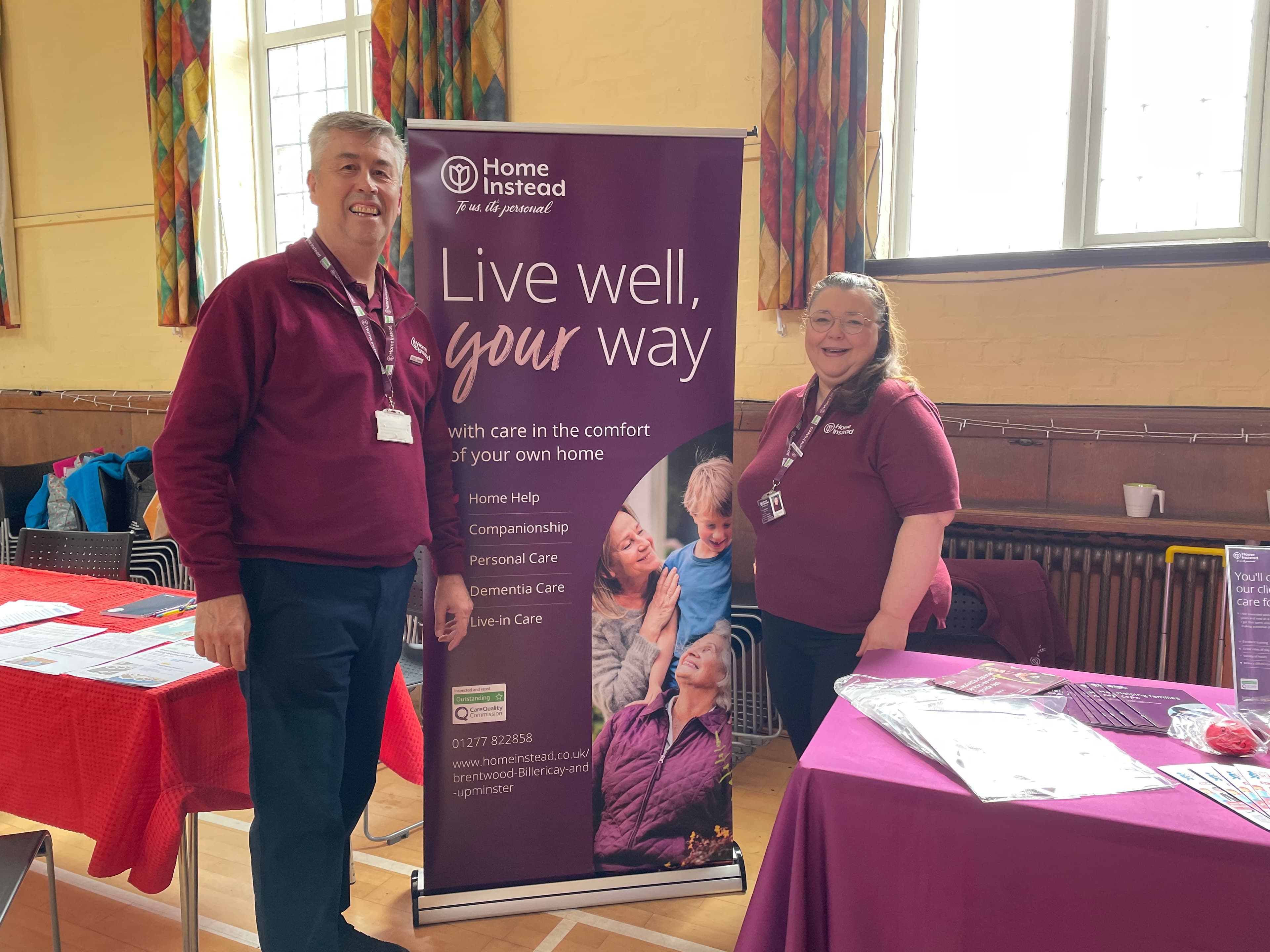 Two people stand by a “Home Instead” banner promoting home care services at an indoor community event. - Home Instead