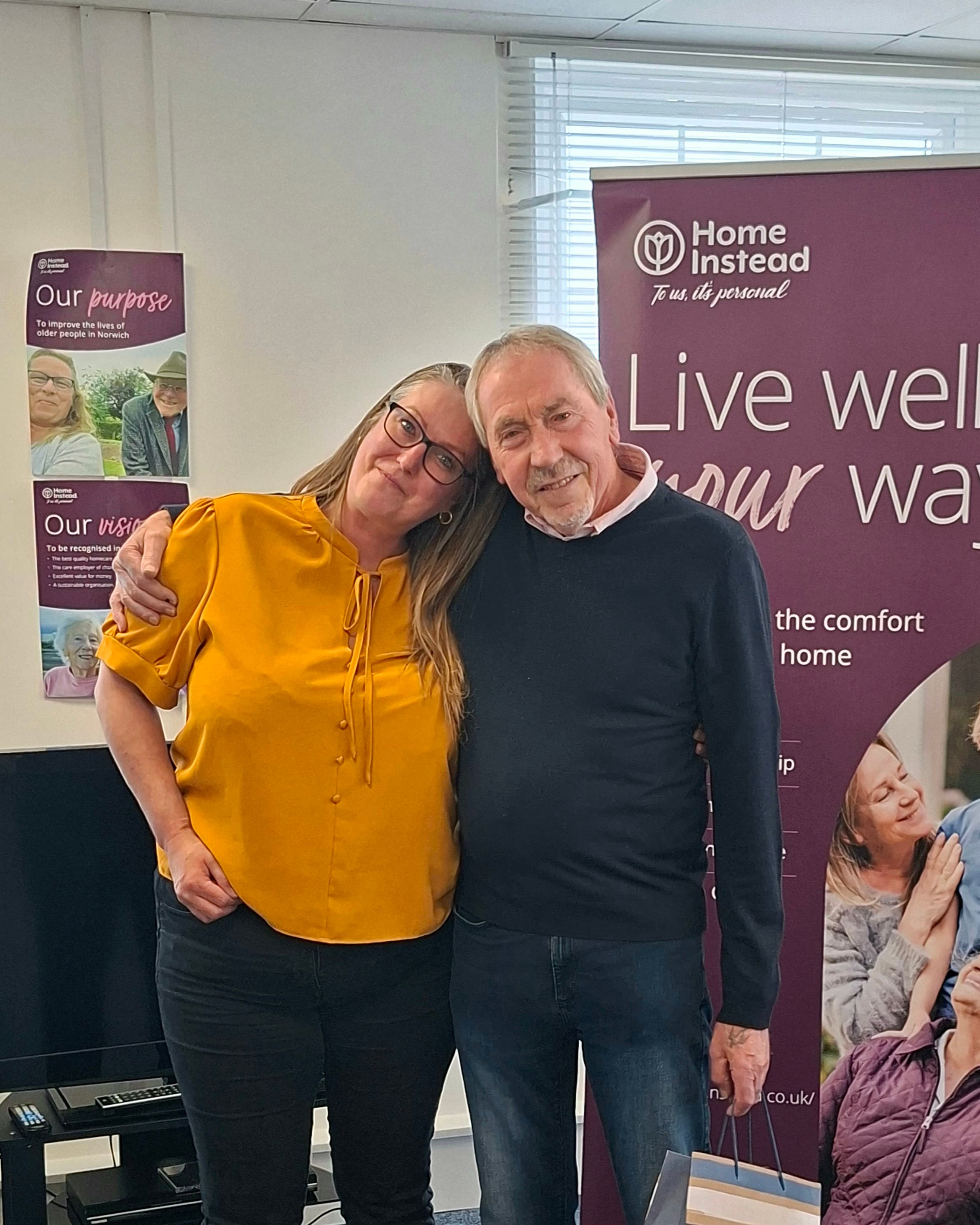 Two people smiling and standing together in front of a "Home Instead" banner in a bright room. - Home Instead