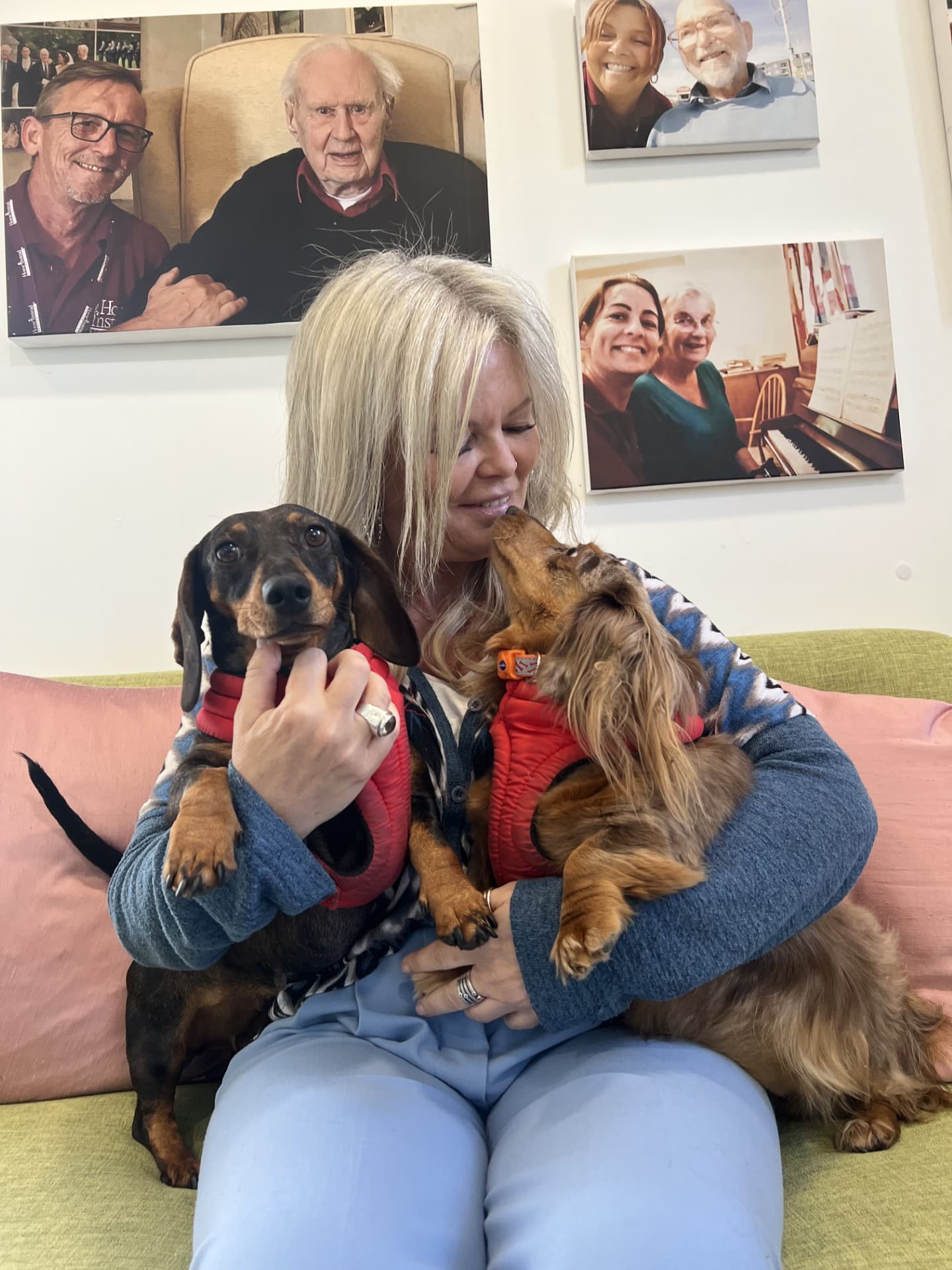 Smiling woman on a couch holds two dachshunds in red harnesses, with photos of people on the wall behind her. - Home Instead