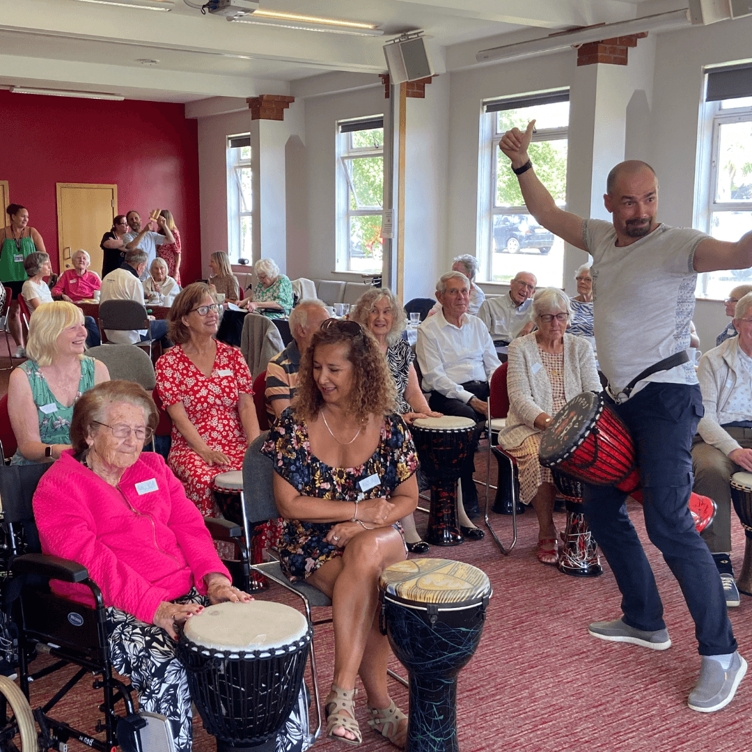 A lively man leads a drumming session with a group of smiling older adults in a bright room. - Home Instead
