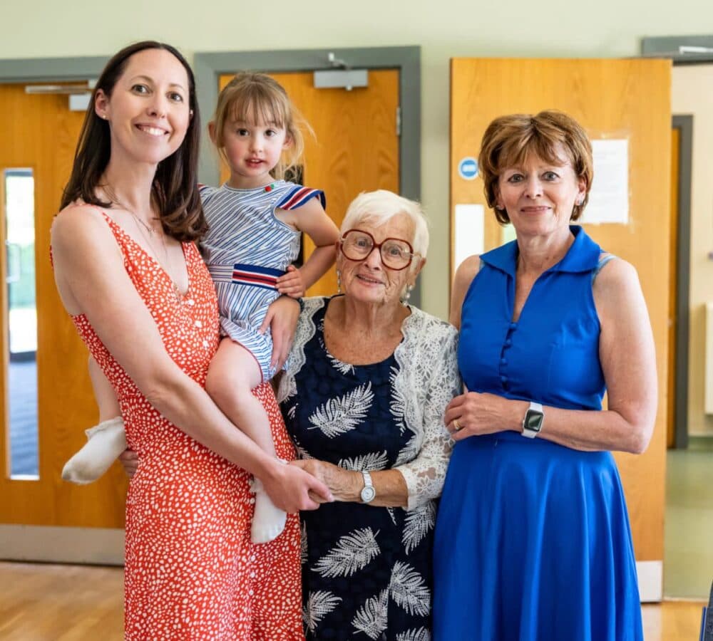 Four generations of women posing together indoors, all smiling at the camera. - Home Instead