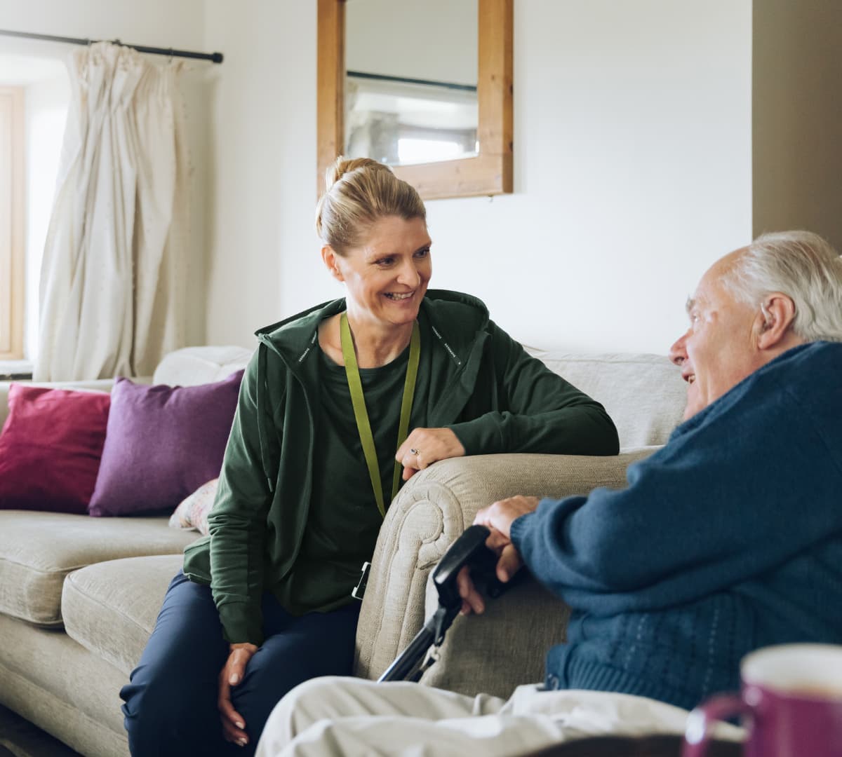 An older male adult with white hair and wearing blue sweater with his younger female carer wearing green and both sitting on a couch while chatting and smiling