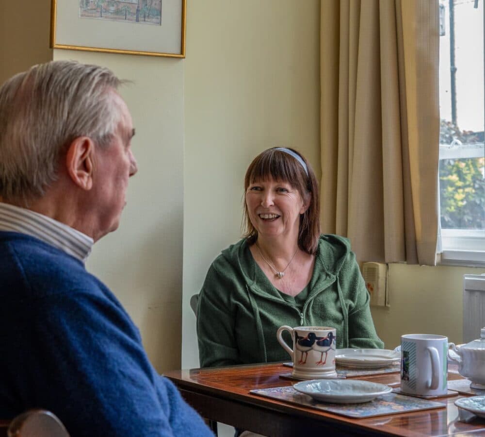 An older male adult with grey hair and wearing eyeglasses while having tea with his younger female carer with short hair and wearing green smiling and happy