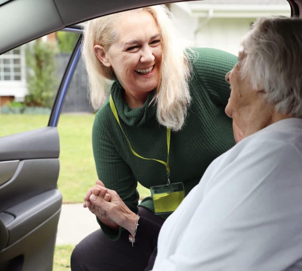 An older female adult in the car while being assisted by her younger female carer with long hair and wearing green both happy and smiling