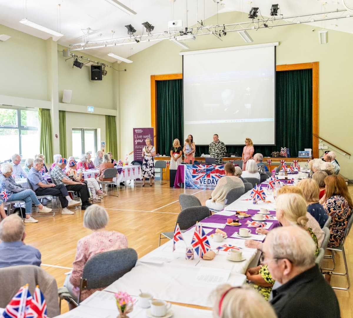 People at tables with Union Jack flags attend an event in a hall, with speakers on stage. - Home Instead