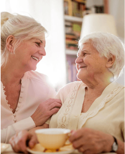 Two women, one younger and one older, smiling at each other while sitting together and enjoying tea. - Home Instead