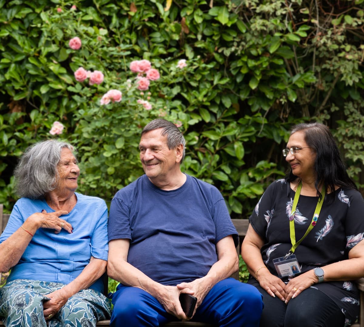 Three older adults sit on a bench, smiling and talking, with green foliage and pink flowers in the background. - Home Instead
