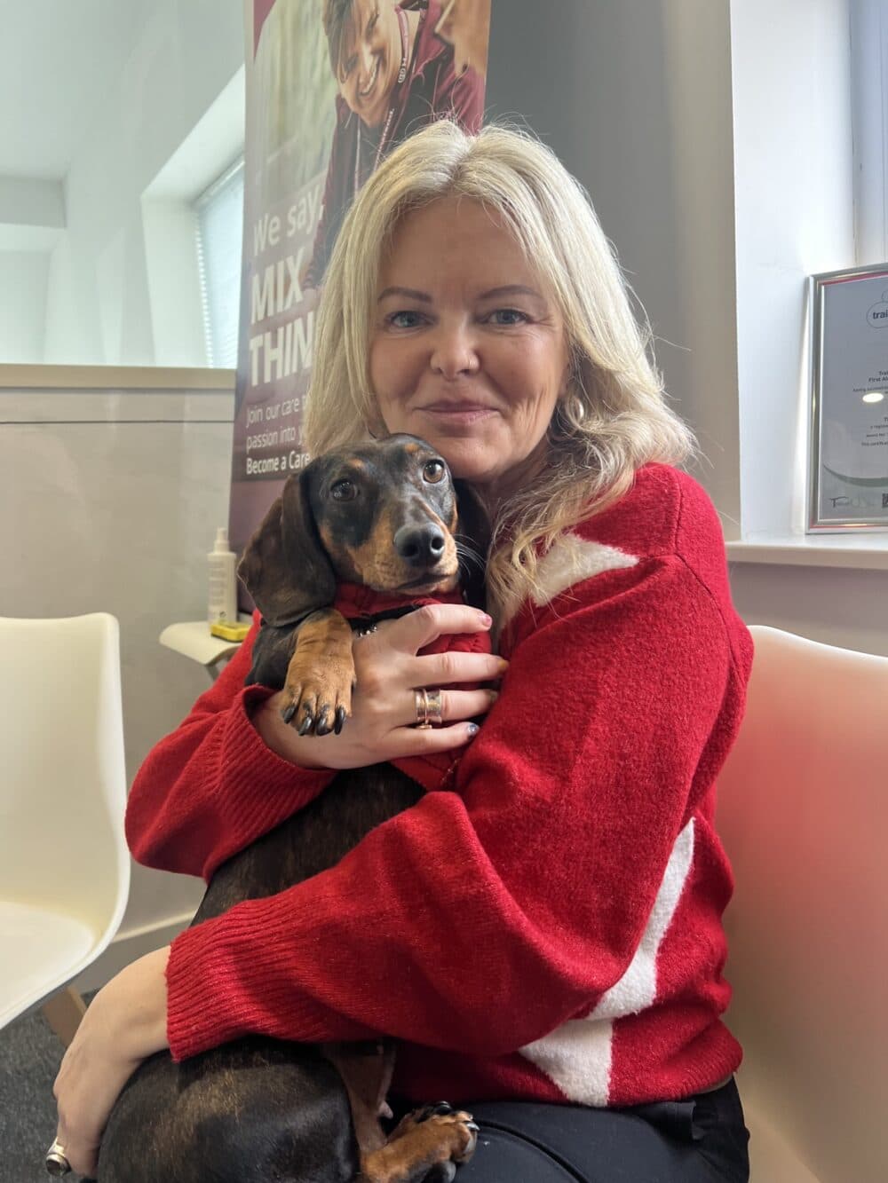 Woman with long blonde hair in a red sweater hugs a dachshund dog while sitting on a chair indoors. - Home Instead