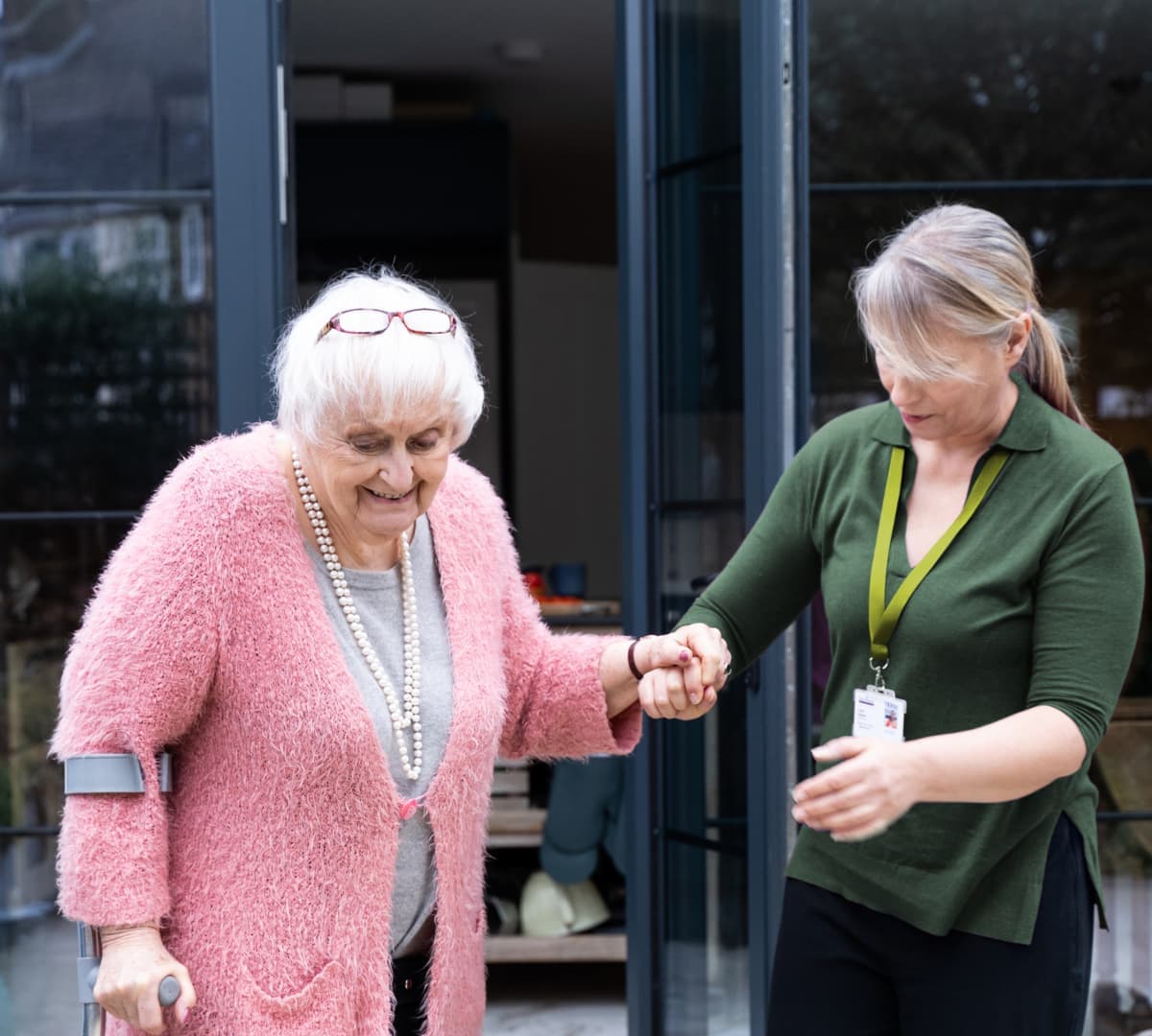 An older female adult with white hair and eyeglasses wearing pink sweater and using a crutch with a younger female carer wearing green