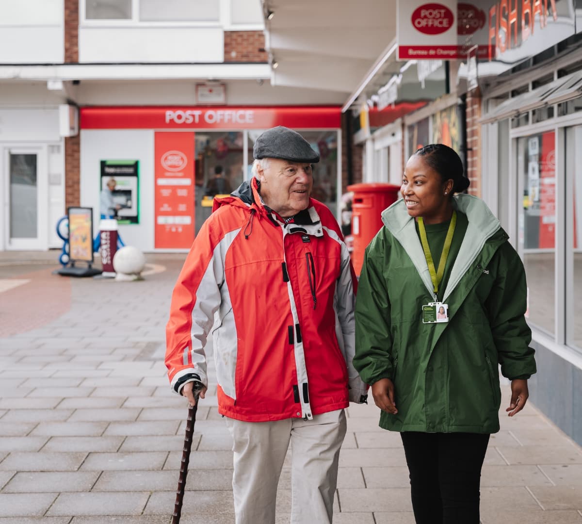 An older male adult wearing cap and with red jacket and using a crane while walking with his younger female carer with green jacket both walking outdoors