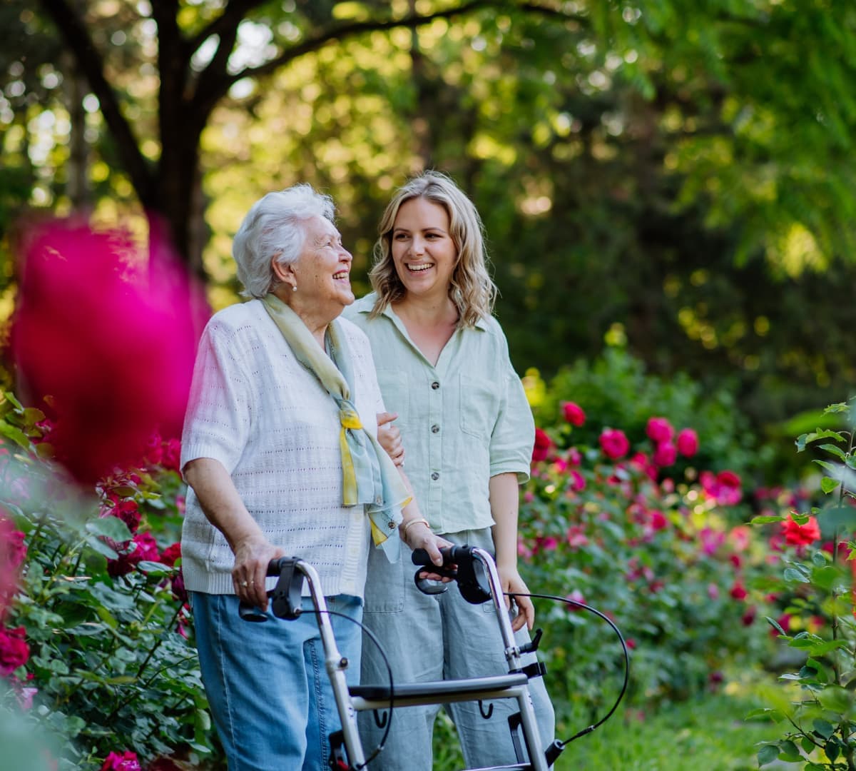 An older female adult with white hair and wearing pink walking at a garden while using a walker with her younger female walker wearing black both happy and smiling and surrounded by green grass